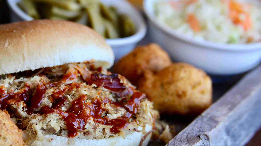 a pulled pork sandwich with hushpuppies, cole slaw and green beans in the background from buddy's bar-b-q in sevierville tn