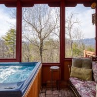 Hot tub on covered porch at Sunny View Cabin with sweeping Smoky Mountain views visible through floor-to-ceiling windows